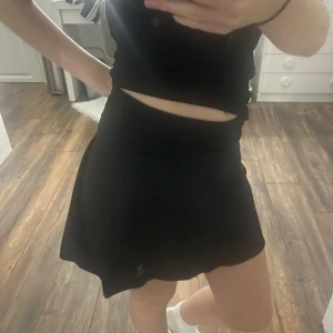 Svart sportkjol från SOC - Svart sportig kortkjol från SOC med inbyggda shorts under. Kjolen har bred midja och är gjord i ett lätt och stretchigt syntetmaterial som andas, perfekt för aktiva dagar. Enkel design utan mönster eller extra detaljer.