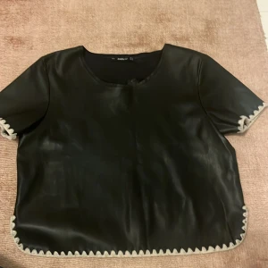 Svart topp i skinn från Zara - Cool svart topp från Zara med skinnlook och korta ärmar. Toppen har beige snörningsdetaljer längs kanterna som ger en unik touch. Rund halsringning och loose passform gör den enkel att styla. Så fin!