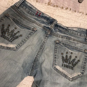 Ljusblå jeans med nitar på bakfickor - Snygga ljusblå jeans med slitningar och raka ben. Bakfickorna är dekorerade med coola nitar i kronmönster som ger extra edge. Jeansen har klassisk femficksdesign, dragkedja och knapp. Perfekta för dig som vill sticka ut lite extra. Det står att dom är L men jag skulle säga M.