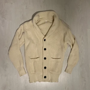 Beige stickad kofta med stora knappar - Säljer en beige stickad kofta med grov struktur och stora svarta knappar framtill. Koftan har två rejäla fickor, bred krage och långa ärmar med ribbade muddar. Perfekt för lager-på-lager och chill stil.