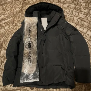 Canada Goose (Black Label) - Svart dunjacka från Canada Goose med huva och avtagbar pälsdetalj. Jackan har diskret logga på ärmen, dragkedja och knappar framtill samt flera fickor. Tillverkad i slitstark polyester och fodrad med dun och fjädrar för extra värme. Perfekt för kalla vinterdagar. (finns i också i stl L)