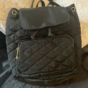 Svart quiltad ryggsäck med snörning - Snygg svart ryggsäck med quiltad design och guldfärgade detaljer. Väskan har en praktisk framficka med dragkedja och snörning upptill under locket. Perfekt storlek för att få plats med allt du behöver och passar till många olika stilar.