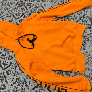 Orange hoodie med svart snörning - Snygg orange hoodie med svart snörning och tryck längs ärmarna. Tröjan har huva och ribbade muddar vid ärmslut och nederkant. Perfekt för dig som gillar färgstarka plagg och streetwear-vibe.