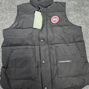 Svart dunväst från Canada Goose - Svart dunväst från Canada Goose med klassisk logga på bröstet. Västen har två stora fickor framtill med lock och tryckknappar, samt hög krage. Materialet är slitstark polyester och dunfyllning som håller dig varm. Perfekt för kalla dagar och snygg att styla över hoodie.