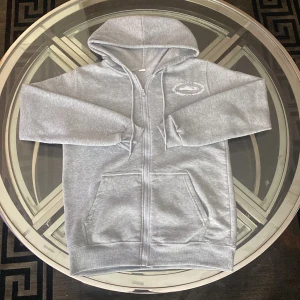 Grå Corteiz zip hoodie med huva - Säljer en grå zip hoodie från Corteiz med stor logga på ryggen och mindre tryck på bröstet. Tröjan har huva, två fickor fram och ribbade muddar. Perfekt för chill dagar och streetwear vibes.