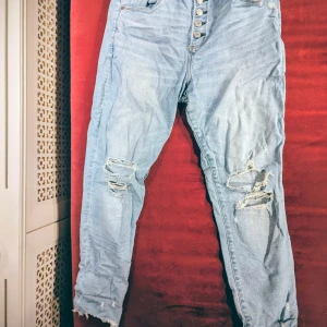 Abercrombie & Fitch Ljusblå ripped jeans med knappar  - A&F high rise super skinny ankle jeans. Säljer ett par ljusblå jeans med slitna detaljer och flera hål på benen. Byxorna har hög midja och knäppning med synliga knappar framtill. Klassisk femficksmodell och raka ben med fransad nederkant. Perfekt för dig som gillar en edgy streetstil.