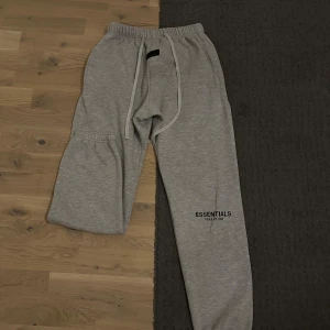 Grå Essentials Fear of God mjukisbyxor - Snygga grå mjukisbyxor från Essentials Fear of God med tryck på benet. Byxorna har elastisk midja med snörning, ribbade muddar vid benslut och en bakficka. Perfekta för chill eller streetwear-stil.