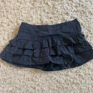 Mörkblå volangkjol från Bershka - Supersöt mörkblå kortkjol från Bershka med flera lager volanger och råa kanter för en edgy look. Kjolen har dragkedja i sidan och är gjord i bomullsmix som känns lätt och skön. Perfekt för dig som gillar en trendig och lekfull stil. Den har inbyggda shorts. 