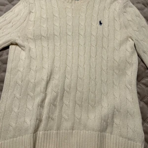 Vit stickad tröja från Polo Ralph Lauren - Säljer en klassisk vit kabelstickad tröja från Polo Ralph Lauren i storlek M. Tröjan har rund hals, ribbade muddar och det ikoniska blå broderade logot på bröstet. Perfekt för dig som gillar stilrena och tidlösa plagg.