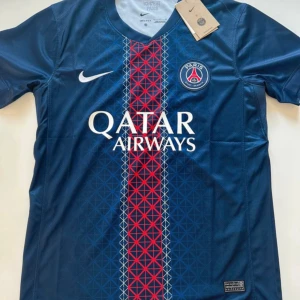 PSG matchtröja Nike blå röd - Snygg PSG fotbollströja från Nike i mörkblått med röd och vit rand längs mitten, dekorerad med geometriskt mönster. Tröjan har klubbmärke på bröstet och Qatar Airways-tryck. Tillverkad i lätt och ventilerande material, perfekt för match eller träning.