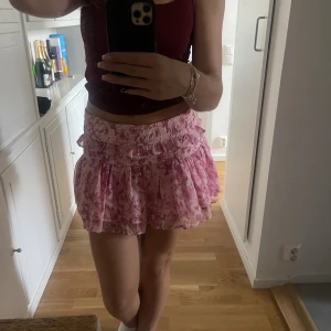 Rosa blommig volangkjol - Supersöt kort kjol i rosa med blommigt mönster och volangdetaljer. Kjolen har en bred, smockad midja som sitter bekvämt och ger en snygg siluett. Perfekt för dig som gillar romantisk och feminin stil.