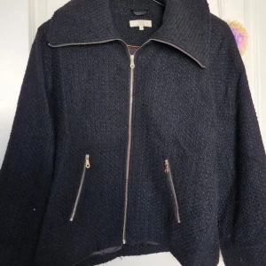 Svart ulljacka med dragkedja från 365 - Svart jacka från 365 med grovstickad yta och bred krage. Jackan har två snedställda fickor med dragkedja och hel dragkedja framtill. Fodrad insida i satin och långärmad modell. Perfekt för dig som gillar stilren och enkel design.