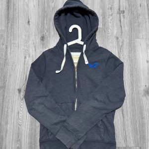 Mörkblå hoodie från Hollister - Snygg mörkblå hoodie från Hollister med dragkedja och vit snörning i huvan. Tröjan har ett blått Hollister-logo på bröstet och praktiska fickor framtill. Perfekt för chill dagar och enkel att matcha med jeans eller joggers.