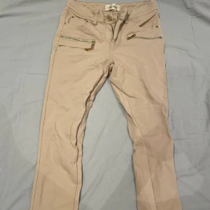 Beiga byxor med guldiga dragkedjor - Snygga beiga byxor med raka ben och coola guldiga dragkedjor framtill. Byxorna har bälteshällor, två fickor bak. Perfekta för en trendig och avslappnad look.