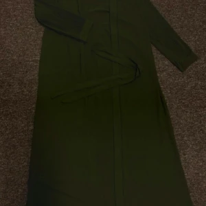 Olivgrön långklänning/cardigan med bälte - Snygg olivgrön långklänning med plisserade långa ärmar och matchande bälte i midjan. Klänningen har en stilren look och är tillverkad i ett mjukt tyg som faller fint. Perfekt för dig som gillar minimalistisk och trendig stil.