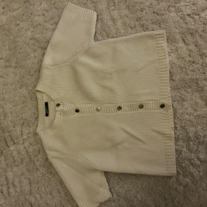 Kortärmad beige kofta från Gina Tricot - Säljer en beige, ribbstickad kofta från Gina Tricot i storlek S. Koftan har korta ärmar och silverfärgade knappar framtill. Den är gjord i ett mjukt stickat material och har en rund halsringning med breda muddar. Perfekt för lager-på-lager och chill stil.