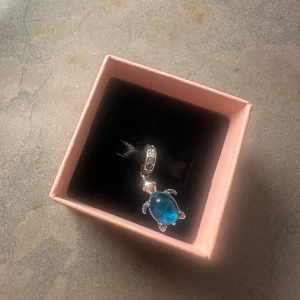 Blått sköldpaddshänge i silver - Supersött hänge i form av en sköldpadda med blå kropp och silverfärgade detaljer. Perfekt att sätta på ett armband eller halsband för en personlig touch. Kommer i en rosa ask och har glittriga stenar på fästet. Perfekt för dig som älskar havsinspirerade smycken.