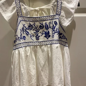 Vit och blå broderad topp - Superfin vit topp med blå broderade blommor och detaljer. Den har en knytning i ryggen och volangiga ärmar. Perfekt på sommaren!