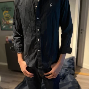 Svart skjorta från Ralph Lauren - Klassisk svart skjorta från Ralph Lauren med vit broderad logga på bröstet. Skjortan har lång ärm, knappar framtill och är tillverkad i bomull. Perfekt för en clean och stilren look. Modellen är 182 men som ser är den lite kort vid längden och ärmarna så skulle passa nån som är 175, passar som en S men igenkligen medium