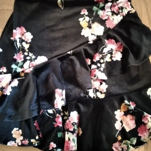 Blommig volangkjol i sammet - Supersöt svart kjol i mjuk sammet med blommigt mönster i rosa, vitt och grönt. Kjolen har volangdetaljer och omlottkänsla som ger en extra snygg look. Perfekt för dig som vill ha något unikt och trendigt!