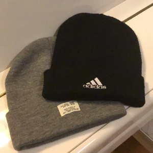 Svart Adidas mössa och grå mössa - Två stilrena mössor, en svart från Adidas med broderad vit logga framtill och en enkel grå mössa utan synligt märke. Båda är klassiskt ribbstickade och perfekta för kyliga dagar. 25krst