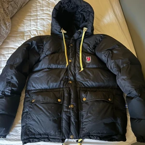Svart pufferjacka från Fjällräven - Säljer en svart pufferjacka från Fjällräven med huva och gula snören. Jackan har broderad logga på bröstet, stora fickor med knappar och dragkedja framtill. Perfekt för kalla dagar. Säljer då den börjar bli för liten, jackan är nytvättad och torktumlad med tennisbollar🙂