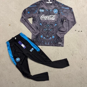 Napoli långärmad fotbollströja EA7 - Säljer en grå och svart långärmad fotbollströja från Napoli med futuristiskt mönster, Coca-Cola tryck och blå detaljer. Märkt med EA7 och Napoli-logga. Materialet är lätt och andas, perfekt för träning eller match.
