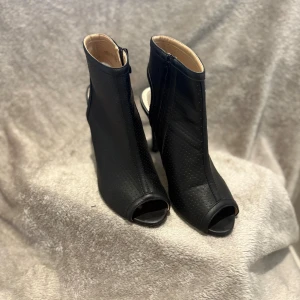 Svarta stövletter med öppen tå och klack - Snygga svarta stövletter med hög smal klack och öppen tå. Skorna är i perforerat syntetmaterial och har dragkedja på sidan för enkel påtagning. Perfekta för dig som vill ha en edgy och trendig look.