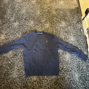 Marinblå kabelstickad tröja Polo Ralph Lauren - Snygg marinblå kabelstickad tröja från Polo Ralph Lauren i 100% bomull. Klassisk rund hals och långärmad modell med röd broderad logga på bröstet. Perfekt för dig som gillar stilrena och tidlösa plagg med premiumkänsla.
