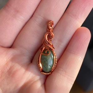 Handgjord berlock med grön sten - Unik berlock med en oval grön sten infattad i snirklig koppartråd. Hängsmycket har en cool, bohemisk vibe med detaljerade tvinnade och virade mönster. Perfekt för dig som gillar naturinspirerade accessoarer och vill sticka ut med något personligt.