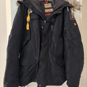 Svart Parajumpers parkajacka XL - Svart parkajacka från Parajumpers med päls på huvan, flera fickor, gul detaljrem och patch på ärmen. Jackan har dragkedja och knappar framtill, samt slitstarka material och slim fit. Perfekt för kalla dagar med snygg streetstil.