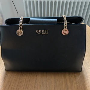 Svart handväska från Guess med guldkedja - Snygg svart handväska från Guess med guldfärgade kedjehandtag och logga framtill. Väskan har flera fack, dragkedja och är tillverkad i slätt skinnliknande material. Perfekt storlek för att få plats med allt du behöver och ger en lyxig känsla till din outfit.