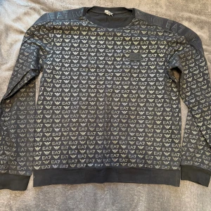 Svart EA7 Emporio Armani sweatshirt - Svart sweatshirt från EA7 Emporio Armani med all-over logomönster i silver. Tröjan har rund hals, ribbade muddar och lång ärm. Snygg och streetig stil som sticker ut med sitt tydliga logotryck.