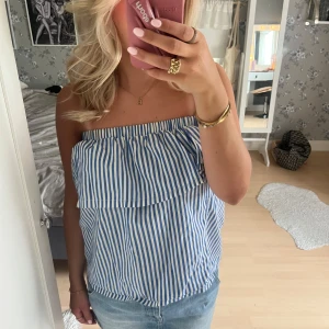 Blåvit randig axelbandslös topp - Säljer en blåvit axelbandslös topp med raka ränder och volangdetalj upptill. Toppen har en luftig passform och är perfekt för varma dagar. Snygg till både jeans och kjol!