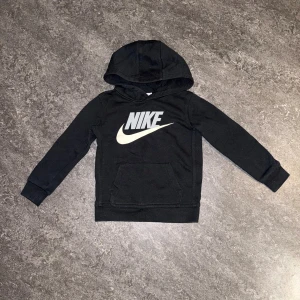 Svart Nike hoodie med logga - Svart hoodie från Nike med stor vit logga på bröstet. Tröjan har huva, lång ärm och en klassisk magficka framtill. Tillverkad i mjuk bomullsmix som är skön att ha på sig. Perfekt för dig som gillar sportig och enkel stil.