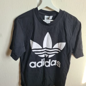 Svart Adidas t-shirt med stor logga XS - Svart t-shirt från Adidas i storlek XS med stor vit Trefoil-logga framtill. Klassisk rund halsringning och korta ärmar. Tillverkad i mjuk bomull som känns skön mot huden. Perfekt för dig som gillar sportig och stilren streetwear.