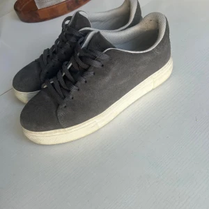 Grå mockasneakers Selected Homme - Snygga sneakers från Selected Homme i mörkgrå mocka med vita sulor och grå snörning. Skorna har en klassisk låg profil och rund tå, perfekt för en clean och stilren look. Insidan är ljusgrå och materialet känns mjukt och bekvämt.
