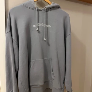 Grå/blå hoodie från Dr Denim med tryck - Säljer en grå/ blå hoodie från Dr Denim i storlek M. Tröjan har huva med snörning, magficka och ett litet vitt tryck på bröstet. Materialet är mjukt och fleecefodrat på insidan, perfekt för chill dagar. Passformen är loose och ärmarna är långärmade. Den är även Oversights 