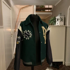 Grön och marinblå bomberjacka  - Snygg bomberjacka i mörkgrönt och marinblått med vita detaljer. Jackan har broderade patches med text och basebollmotiv, samt NY-logga på bröstet. Ribbad mudd vid ärmslut och nederkant, knappar framtill och kontrastfärgade ärmar. Perfekt för dig som gillar sportig streetstyle.