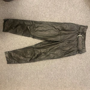 Svarta skinnbyxor med bälte - Snygga svarta skinnbyxor med tillhörande bälte och raka ben. Byxorna har hög midja och en stilren look som passar perfekt till en trendig outfit. Materialet är skinnimitation och ger en cool vibe. Perfekta för dig som vill sticka ut lite extra.