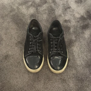 Svarta Lanvins - Svarta sneakers med snygg lackad tå och vit sula. Skorna har snörning och en klassisk låg profil. Ovandelen är i textil och tån är i blankt material som ger en cool kontrast. Perfekta för dig som gillar stilrena men ändå unika sneakers.