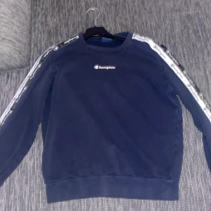 Blå sweatshirt från Champion - Mörkblå sweatshirt från Champion med logga på bröstet och vita band med Champion-tryck längs ärmarna. Klassisk rund hals och långa ärmar. Perfekt för en sportig och avslappnad stil.