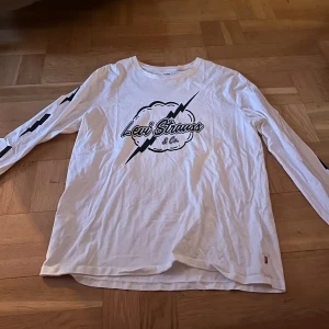 Vit långärmad Levi's tröja med tryck - Vit långärmad tröja från Levi's med svart blixt- och molntryck på bröstet samt svarta blixtar längs ärmarna. Använd ett par gånger.
