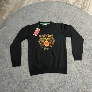 Svart sweatshirt med tigerbrodyr - Svart sweatshirt med ikonisk tigerbrodyr i gult och rosa på bröstet. Tröjan har rund hals, ribbade muddar och klassisk passform. Perfekt statement-plagg för dig som gillar streetstyle och vill sticka ut.