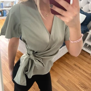 Ljusgrön omlottblus med knyt - Snygg ljusgrön omlottblus med v-ringning och korta, vida ärmar. Blusen har ett brett knytband i midjan som ger en fin siluett. Perfekt att styla med både jeans och kostymbyxor för en trendig look.