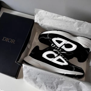 Dior B30 svarta och vita sneakers - Säljer ett par Dior B30 sneakers i svart och vitt med tydlig Dior-logga på sidan. Skorna har en chunky vit sula, svarta mesh- och skinnpaneler samt vita snören. Snygg och modern siluett som sticker ut, perfekt för dig som gillar exklusiva sneakers.