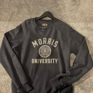 Morris tröja - Blå sweatshirt från Morris med klassiskt universitetsprint i vitt framtill. Tröjan har rund hals, ribbade muddar och lång ärm. Perfekt för dig som gillar college-vibe och vill ha en enkel, stilren look. Materialet är mjukt och skönt, troligen bomull.