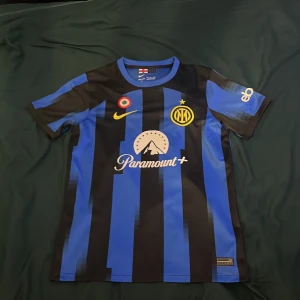 Inter Milan matchtröja Nike S - Inter Milan fotbollströja från Nike i Dri-FIT-material. Klassisk blå och svart randig design med klubbmärke och stjärna på bröstet, samt Paramount+ sponsortryck. Kortärmad modell med modern passform och broderade detaljer.