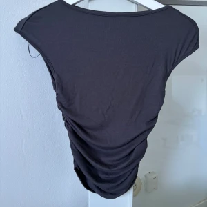 Svart ärmlös croppad topp - Säljer en svart ärmlös topp med croppad passform och snygga rynkade detaljer längs sidorna. Toppen har en enkel, stilren design och är tillverkad i mjukt bomullsmaterial. Perfekt att matcha med höga jeans eller kjol.