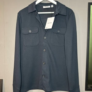Mörkblå overshirt från Only & Sons - Stilren mörkblå overshirt från Only & Sons med klassisk krage, två bröstfickor med knappar och långa ärmar. Overshirten har en clean look och är tillverkad i ett mjukt material som känns skönt mot huden. Perfekt för dig som gillar enkel och snygg stil. Overshirten är helt ny, hör av er vid frågor!☺️